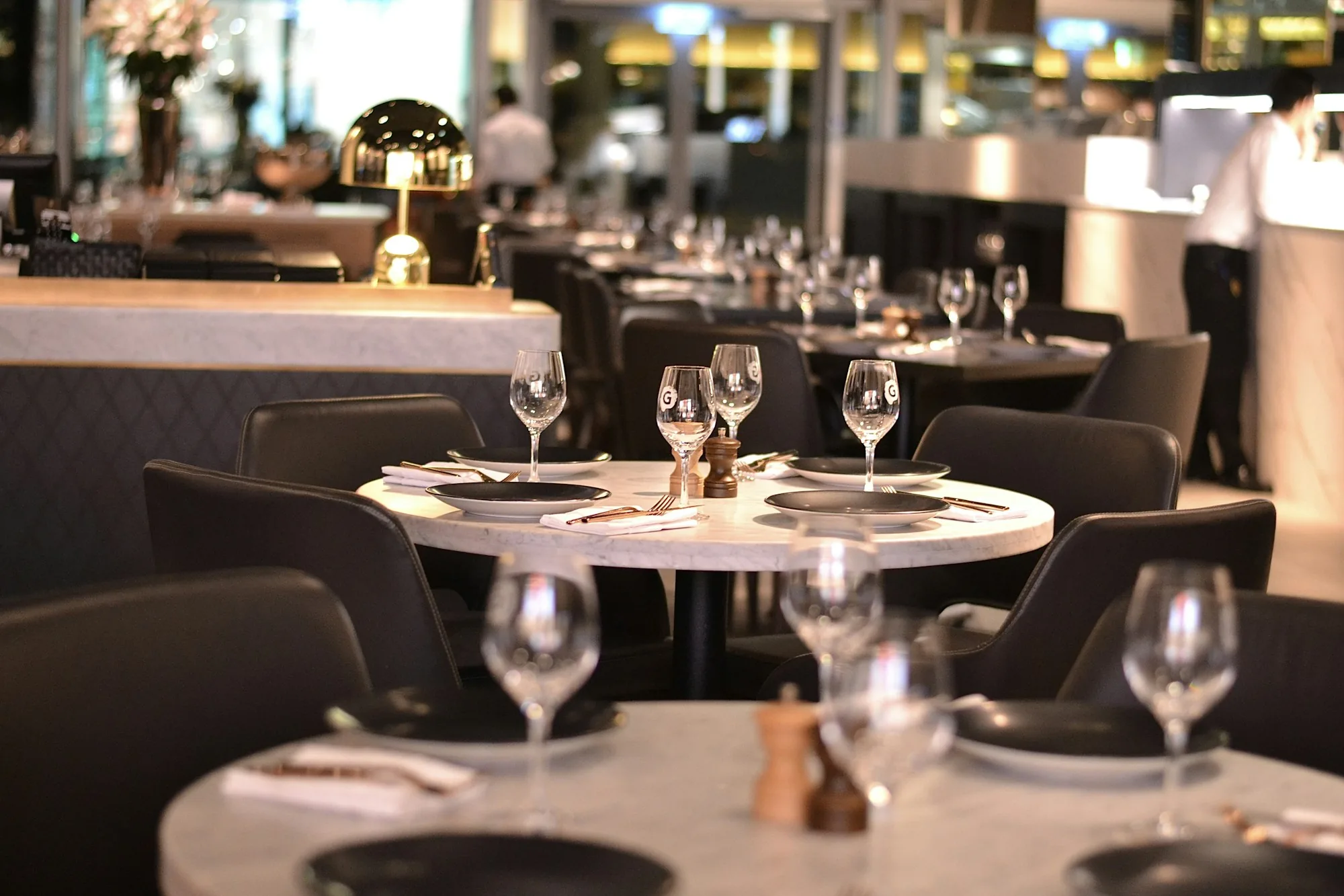 A hip restaurant dining room with marble tables set for dinner and a host stand visible in the background — the reservation moment, ready for service